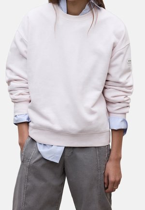Personne portant un sweat-shirt rose clair sur une chemise bleue à col avec un pantalon gris, mains dans les poches, cadré à la poitrine et aux cuisses.