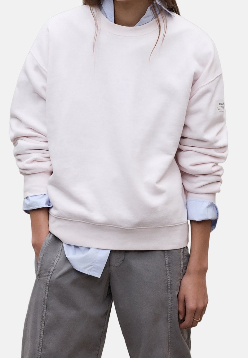 Personne portant un sweat-shirt rose clair sur une chemise bleue à col avec un pantalon gris, mains dans les poches, cadré à la poitrine et aux cuisses.