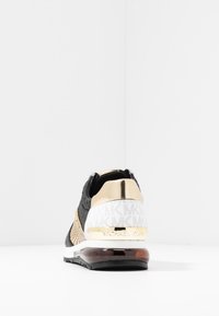 Black, white, and gold sneaker with Michael Kors logo on heel, textured upper, and translucent brown air cushion sole against white background.