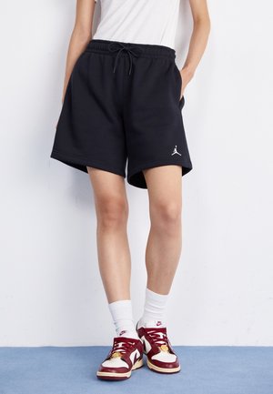 Person iført sorte Jordan-shorts, hvide ankelsokker og røde og hvide Nike-sneakers, der står op ad en hvid væg på blå gulv.