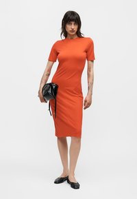 Γυναίκα που φοράει εφαρμοστή πορτοκαλί φόρεμα μήκους μέχρι το γόνατο, κρατώντας μια μαύρη τσάντα, με μαύρα φλατ παπούτσια, στέκεται μπροστά σε ένα απλό φωτεινό φόντο.
