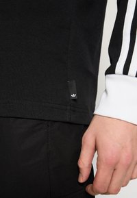 Svart bomullsskjorta med lång ärm och vit manschett, med tre vita ränder på ärmen och en liten svart logotagg vid nederkanten.