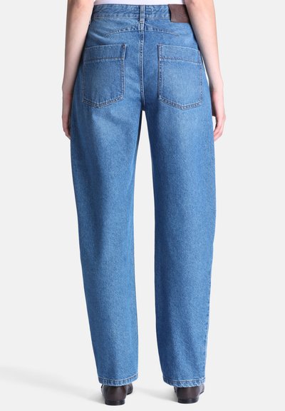 Vue arrière d'une personne portant un jean en denim bleu droit avec des poches arrière et des chaussures marron sur fond blanc.