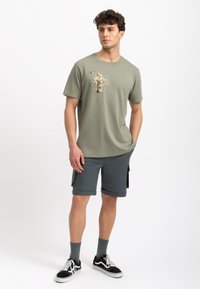T-shirt in cotone verde oliva, maniche corte, con un design stampato sul petto. Abbinato a pantaloni corti grigio scuro fino al ginocchio e sneakers nere.