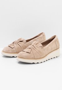 Une paire de mocassins slip-on en suède beige avec un détail de nœud torsadé sur le dessus et des semelles à plateforme blanches à crans.