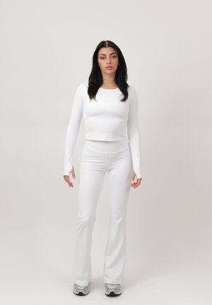 Kvinde med langt mørkt hår iført hvid tætsiddende langærmet top og hvide vide bukser, står foran en ensfarvet lys baggrund.