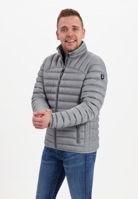 Grå pufferjacka med horisontell sömnad, dragkedja framtill, hög krage och två sidofickor med dragkedja. Bärs tillsammans med blå jeans.