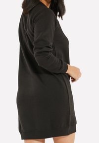 Robe sweat-shirt noire à manches longues, col arrondi et poignets côtelés. Texture douce et coupe décontractée, arrivant à mi-cuisse.