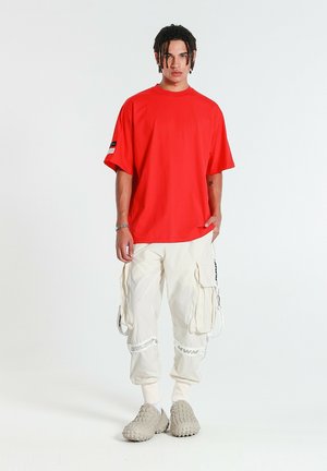 Joven con cabello trenzado viste una camiseta roja holgada, pantalones cargo blancos con tirantes y zapatos beige texturizados sin cordones sobre fondo blanco.