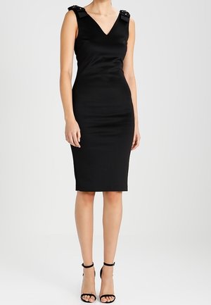 Femme portant une robe noire cintrée sans manches avec des bretelles ornées et des sandales noires à talons hauts, debout sur un fond uni.
