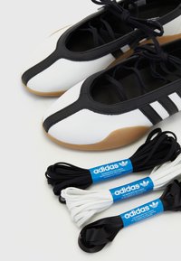 Tênis atléticos brancos e pretos com uma sola de borracha distinta, apresentando três riscas pretas; inclui cordões adicionais pretos e brancos.