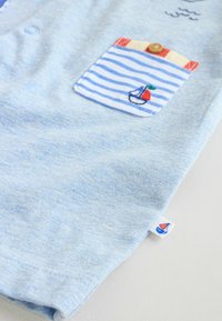 Camiseta azul claro de manga corta con un bolsillo a rayas que presenta un velero y una cereza. Textura de algodón suave, diseño casual y detalle de botón.