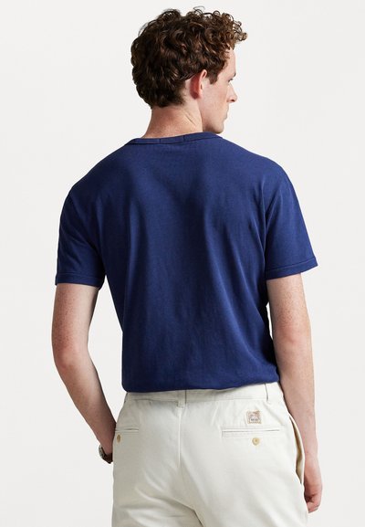 Tricou din bumbac bleu marin, cu mâneci scurte și o croială lejeră, văzut din spate, purtat cu pantaloni crem, care prezintă o etichetă.