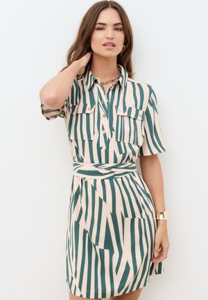 Vrouw in een kort mouws, ceintuurloze jurk met groene en beige verticale zigzagstrepen, staand tegen een witte achtergrond, met haar hand bij haar nek.