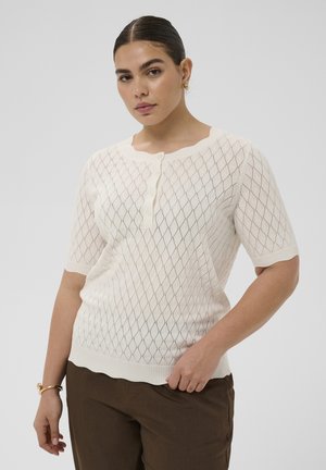 Naine, kellel on tagasiharjatud juuksed, kannab valget lühikeste varrukatega kootud särki teemantmustriga ja pruune pükse, seisab ühevärvilise tausta ees.