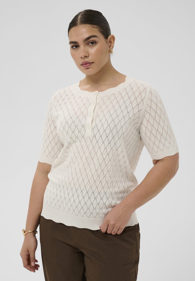 Frau mit zurückgekämmtem Haar, die ein weißes, kurzärmeliges Stricktop mit Rautenmuster und braune Hosen trägt, vor einem einfarbigen Hintergrund stehend.