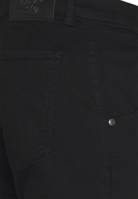Vaqueros de mezclilla negros con una textura suave, que cuentan con un bolsillo trasero, un detalle de botón metálico y una etiqueta de cuero con marca en la cinturilla.