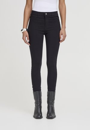 Kvinde iført tætsiddende sorte jeans, sorte læderstøvler til midt på læggen, en hvid ærmeløs top og et sølvarmbånd på hendes venstre håndled.