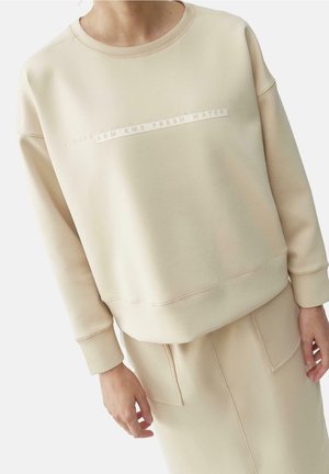 Osoba ubrana w beżową bluzę i pasującą spódnicę z dużymi przednimi kieszeniami, na bluzie napis "35M KM3 WODY ŚWIEŻEJ".