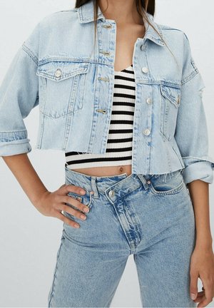 Vrouw draagt een lichtblauw cropped spijkerjasje over een zwart-wit gestreepte crop top met lichtblauwe jeans met hoge taille.