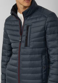 Granatowa kurtka puchowa z pikowanym wzorem, wyposażona w wysoki kołnierz, przedni zamek błyskawiczny oraz kontrastujący czerwony akcent wzdłuż zamka. Posiada kieszeń na piersi.