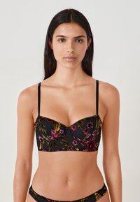 Donna che indossa un set di lingerie ricamato floreale nero con spalline sottili, in piedi contro uno sfondo chiaro e semplice.