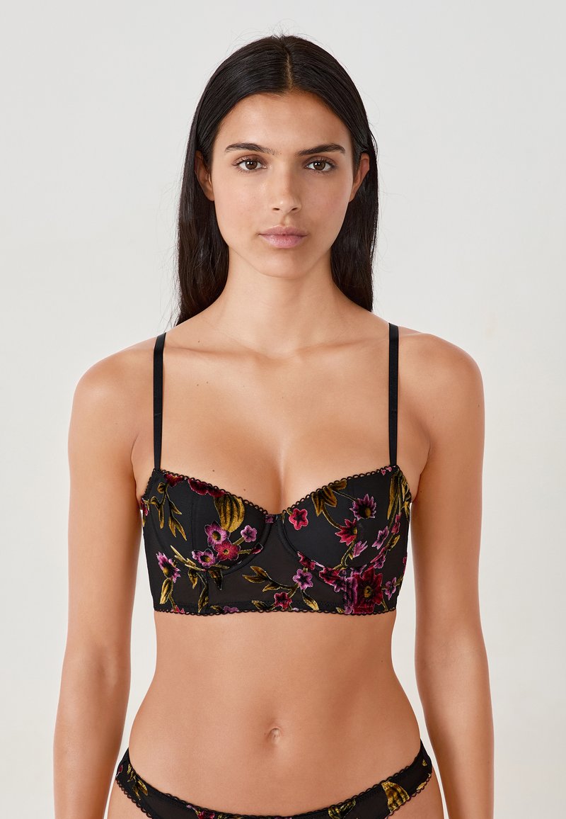 Donna che indossa un set di lingerie ricamato floreale nero con spalline sottili, in piedi contro uno sfondo chiaro e semplice.