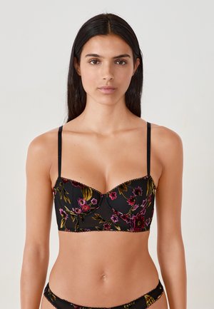 Femme portant un ensemble de lingerie noire brodé de fleurs avec des fines bretelles, se tenant devant un fond clair uni.