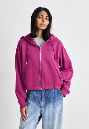 Fuchsia zip-up hoodie with a drawstring hood, front pockets, and ribbed hem. Paired with light blue faded denim pants.