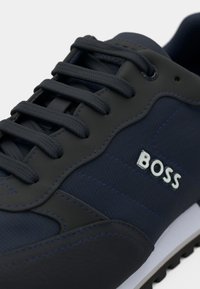 Adidași de culoare bleumarin, cu un material texturat și accente din piele netedă, având șireturi închise la culoare, cusături contrastante și o talpă albă. Logo afisat.