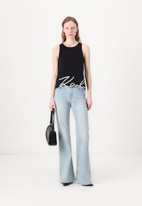Svart ermeløs topp med hvit "Karl" tekst, kombinert med lyseblå utslått jeans. Modellen holder en liten svart veske. Enkel bakgrunn.