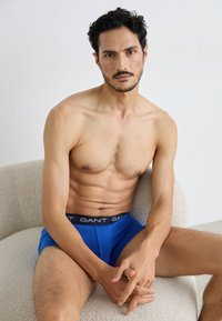 Modré boxerky s čiernym pásom, na ktorom je vyobrazené logo "GANT". Model má štíhlu postavu a sedí na textúrovanej svetlej stoličke.
