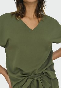 Blusa verde oliva con diseño de cuello en V, mangas cortas y detalle de cintura atada. Fabricada en un tejido ligero y texturizado.