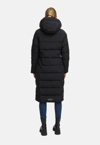 Abrigo de plumas negro con capucha, de diseño largo, con acolchado horizontal y una etiqueta de logo en el dobladillo. Combinado con jeans azules y botas negras.