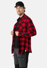 Rood-zwart geruite flanellen shirt, met knoopsluiting, zachte textuur; gedragen over een zwart shirt met grijze broek en zwarte accenten.