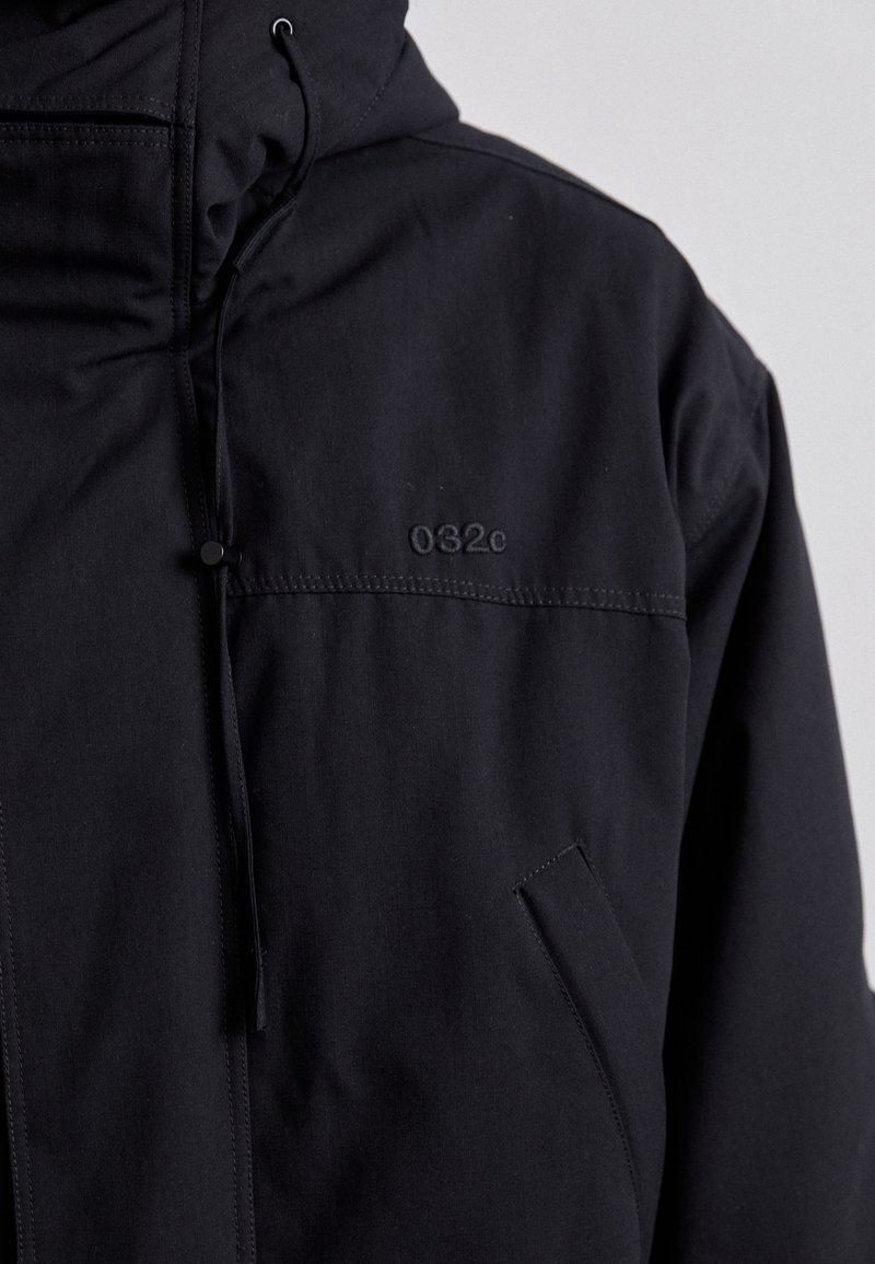 Schwarze Kapuzenjacke aus strukturiertem Stoff mit einer Fronttasche, Zugbändern an der Kapuze und dezent geprägtem Branding "032c."