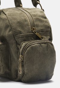 Olive krāsas korduroja soma ar teksturētu ārējo daļu, kas piedāvā galveno rāvējslēdzēja nodalījumu un mazāku priekšējo rāvējslēdzēja kabatu ar misiņa furnitūru.