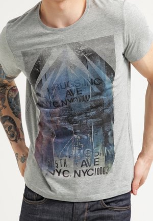 Giovane uomo che indossa una maglietta grigia con un design di paesaggio urbano e testo che mostra "5th Ave NYC" e tatuaggi visibili sul braccio sinistro.