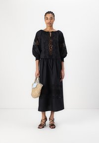 Completo nero a due pezzi composto da una blusa corta con maniche a sbuffo e dettagli ricamati, abbinato a una gonna fluida. Capelli intrecciati, borsa intrecciata.