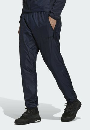 Persona vestida con pantalones deportivos azul oscuro con bolsillos con cremallera y zapatillas de senderismo negras, de pie con una mano en el bolsillo contra un fondo liso.