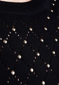 Czarny sweter dzianinowy z wycięciami w kształcie diamentów, ozdobiony okrągłymi metalowymi ćwiekami. Żebrowany dekolt i fakturowana tkanina.