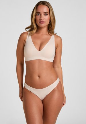 Vrouw met lang blond haar die een beige naadloze beha en bijpassend ondergoed draagt tegen een effen grijze achtergrond.
