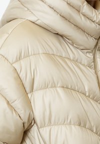 Béžová péřová bunda s prošívaným designem, s hladkým, lesklým povrchem a kapucí. Viditelné švy a detaily švů zvyšují strukturu.