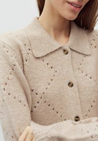 Bézs színű kötött kardigán gallérral, gombokkal, apró gyémánt alakú lyukakkal díszített texturált mintával és bordázott mandzsettákkal.