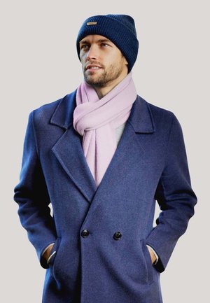 Cappotto di lana blu con due bottoni neri, indossato sopra un maglione grigio. Un sciarpa rosa chiaro a coste e un berretto lavorato a maglia blu navy completano il look.