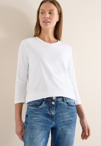 Haut blanc en coton à manches longues avec un col en V, associé à un jean en denim bleu avec une fermeture à deux boutons et un effet délavé subtil.