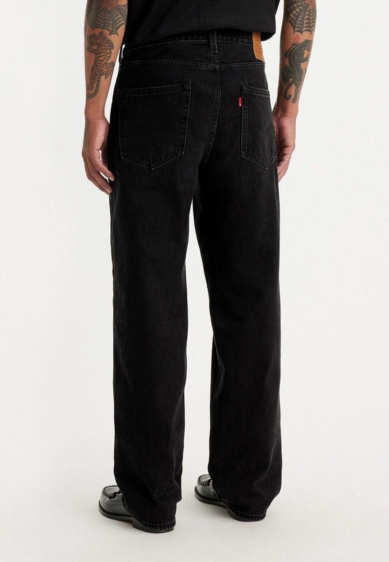 Homme debout portant un jean noir ample et des chaussures noires, avec des bras tatoués visibles de dos sur un fond clair uni.