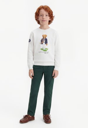 Băiat tânăr cu păr roșcat, purtând un hanorac alb cu un desen cu urs, pantaloni verde închis și pantofi maro cu șireturi, stând pe un fundal alb.