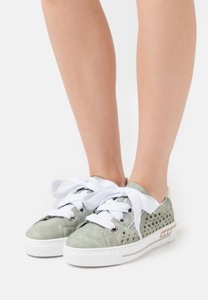 Baskets vert clair avec côtés perforés, lacets blancs épais noués en nœuds, et semelles en caoutchouc blanches portées pieds nus.