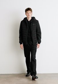 Giacca a vento nera con zip sul davanti e cappuccio, abbinata a pantaloni cargo neri. Entrambi hanno una texture liscia e opaca e dettagli funzionali.